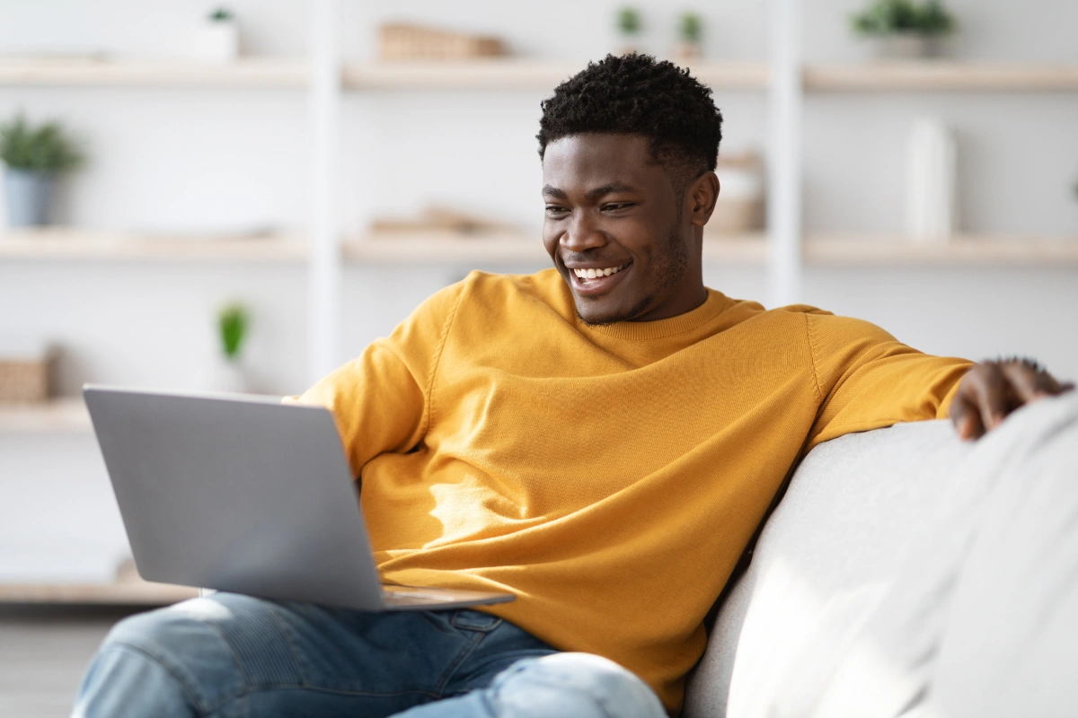 A man on a couch in a yellow sweater smiling and using his laptop in his living room to pay his Frontier internet bill online