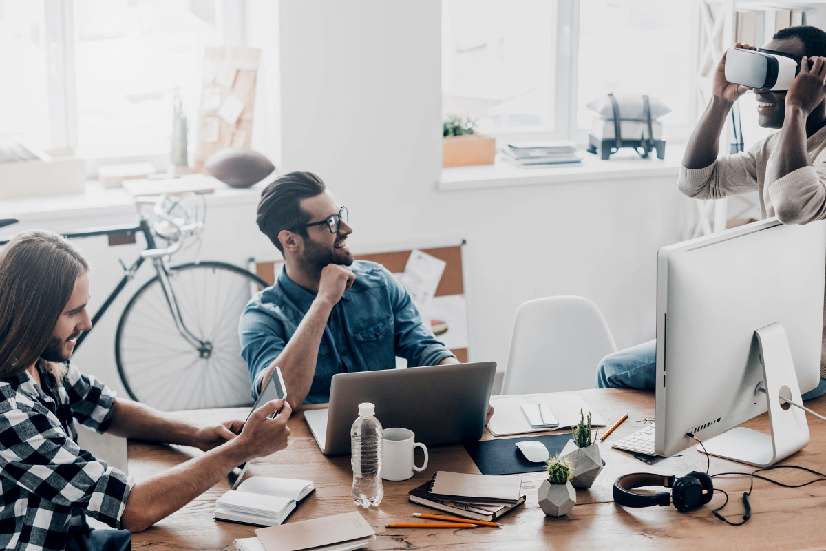A group of co-workers at a desk together, one sitting in front of a laptop, another sitting and using a tablet, and another standing and using a VR headset with Frontier internet
